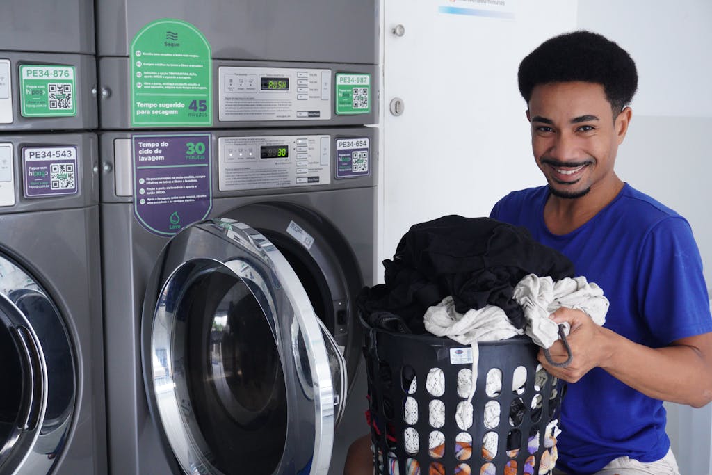 Smiling man holding laundry basket in a laundromat, ready to wash clothes.